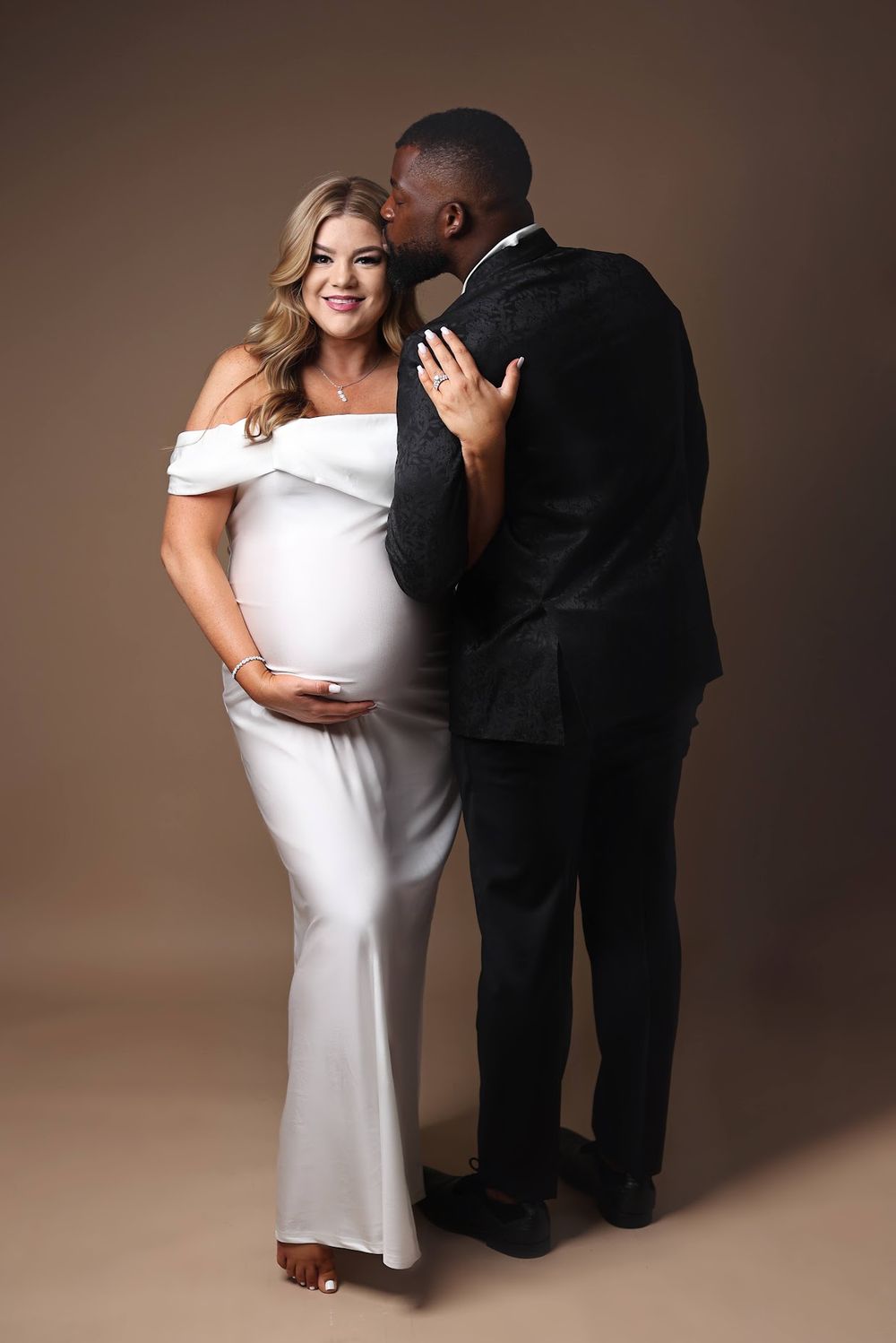 Expecting couple in formal wear, man in black suit kissing pregnant woman in white off-shoulder gown.