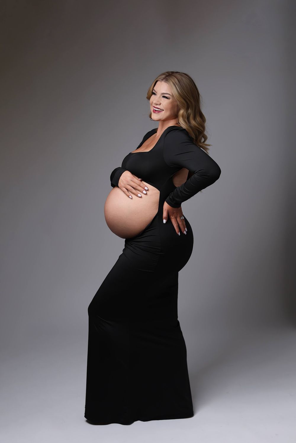 A smiling pregnant woman in a black long-sleeve gown poses sideways, showcasing her baby bump in a studio.