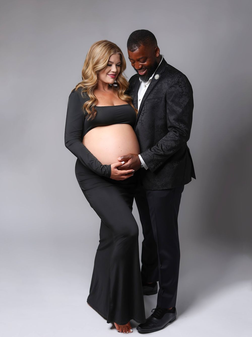 A pregnant woman in black and her partner in a suit tenderly admire her baby bump in a studio portrait.