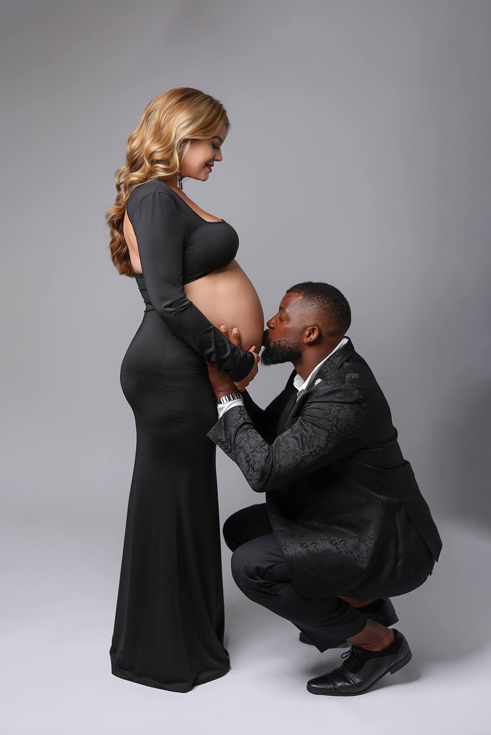 A man in a black suit kneels to kiss a pregnant woman's baby bump during a maternity studio photoshoot.