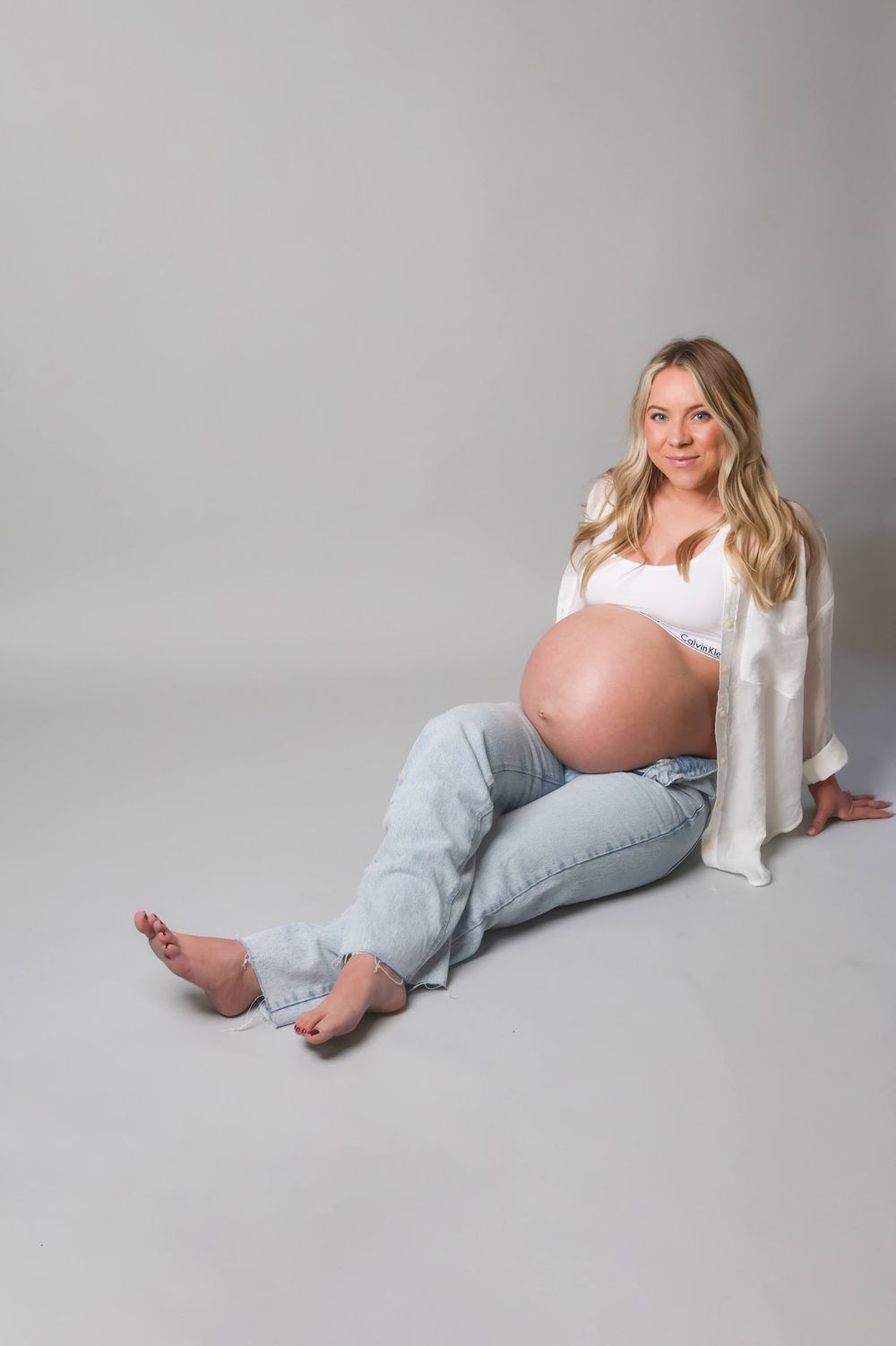 Pregnant woman sitting on floor in light blue jeans and white top, showing bare baby bump in studio setting.