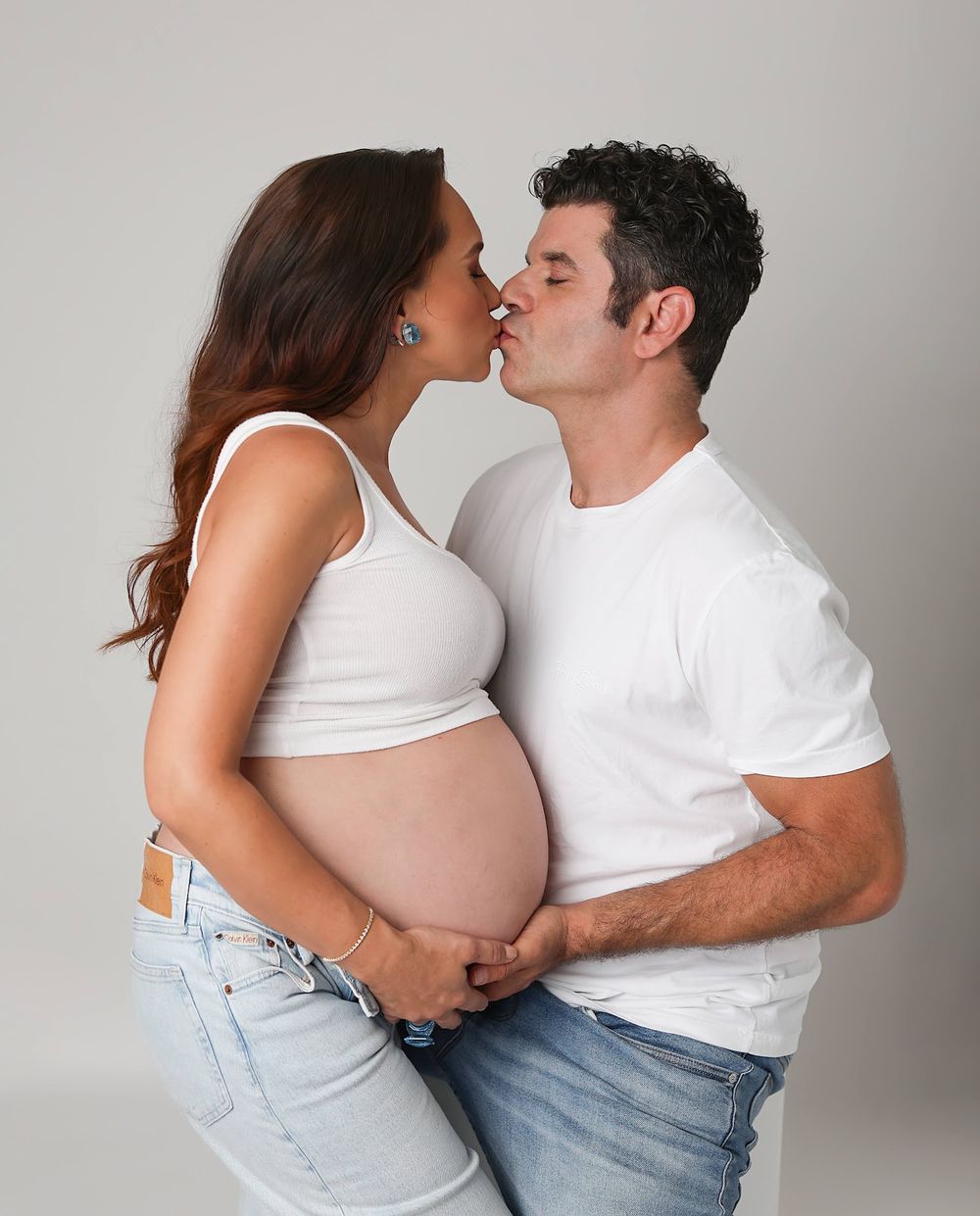 Couple kissing during maternity shoot, man in white tee cradles pregnant womans bare belly in jeans.