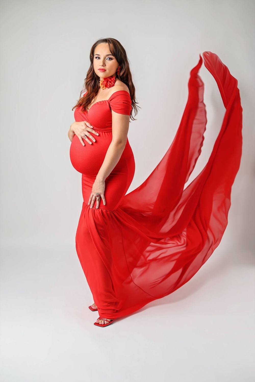 Pregnant woman in flowing red off-shoulder gown with dramatic fabric train in studio maternity shoot.