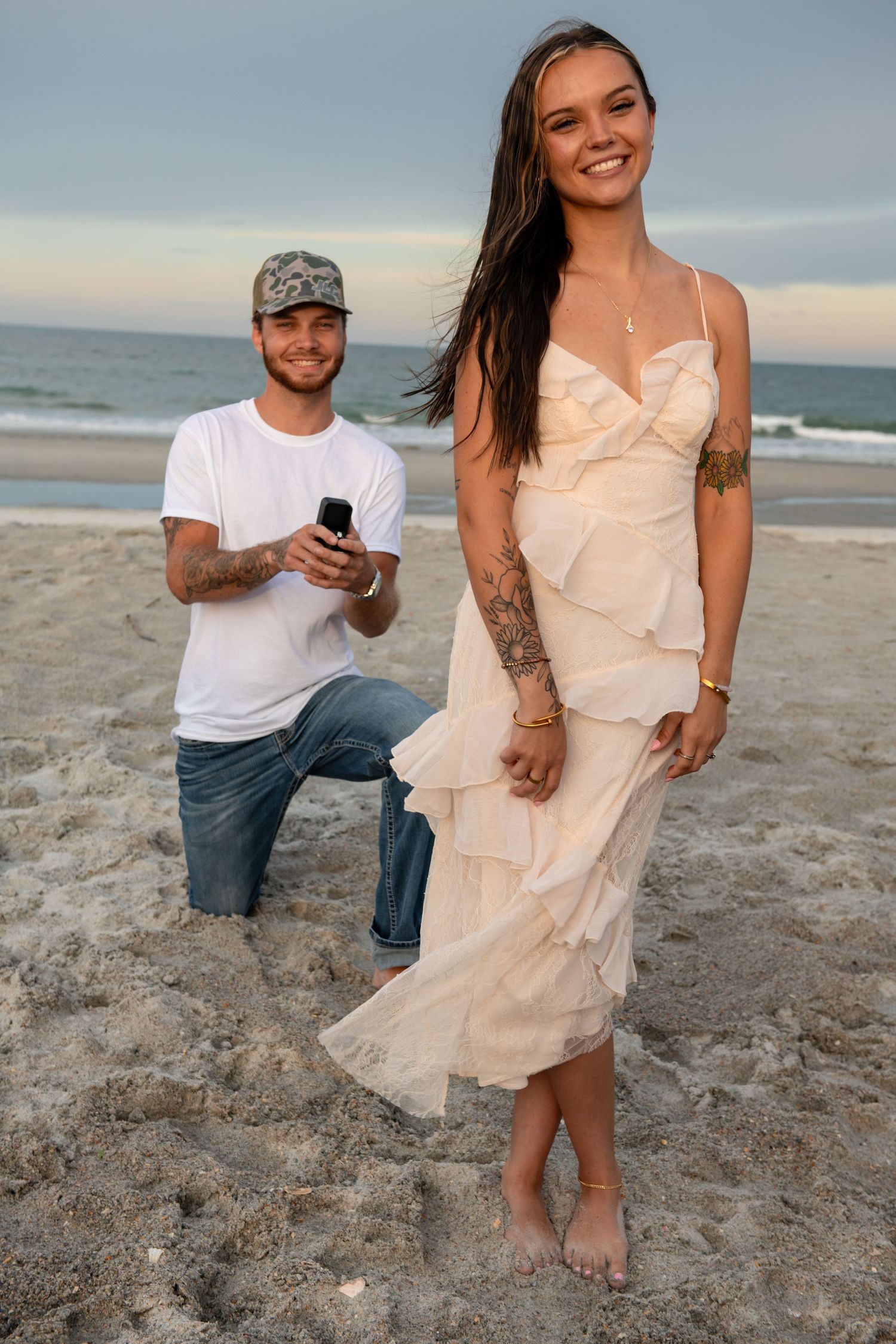 A couple on a beach at sunset, man kneeling with a ring box proposing to a smiling woman in a cream dress.