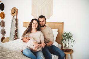 Happy young family with mother, bearded father and newborn baby posing on bed in boho bedroom.