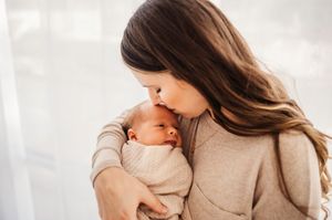 Mother with long brown hair gently kissing her swaddled newborn baby against bright window.