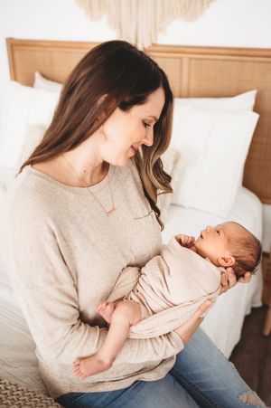 Smiling brunette mother in beige sweater holding and gazing at her newborn baby indoors.
