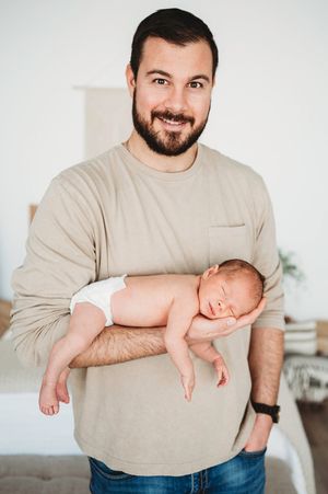 Smiling bearded father in tan shirt proudly holding sleeping newborn baby in bright bedroom.