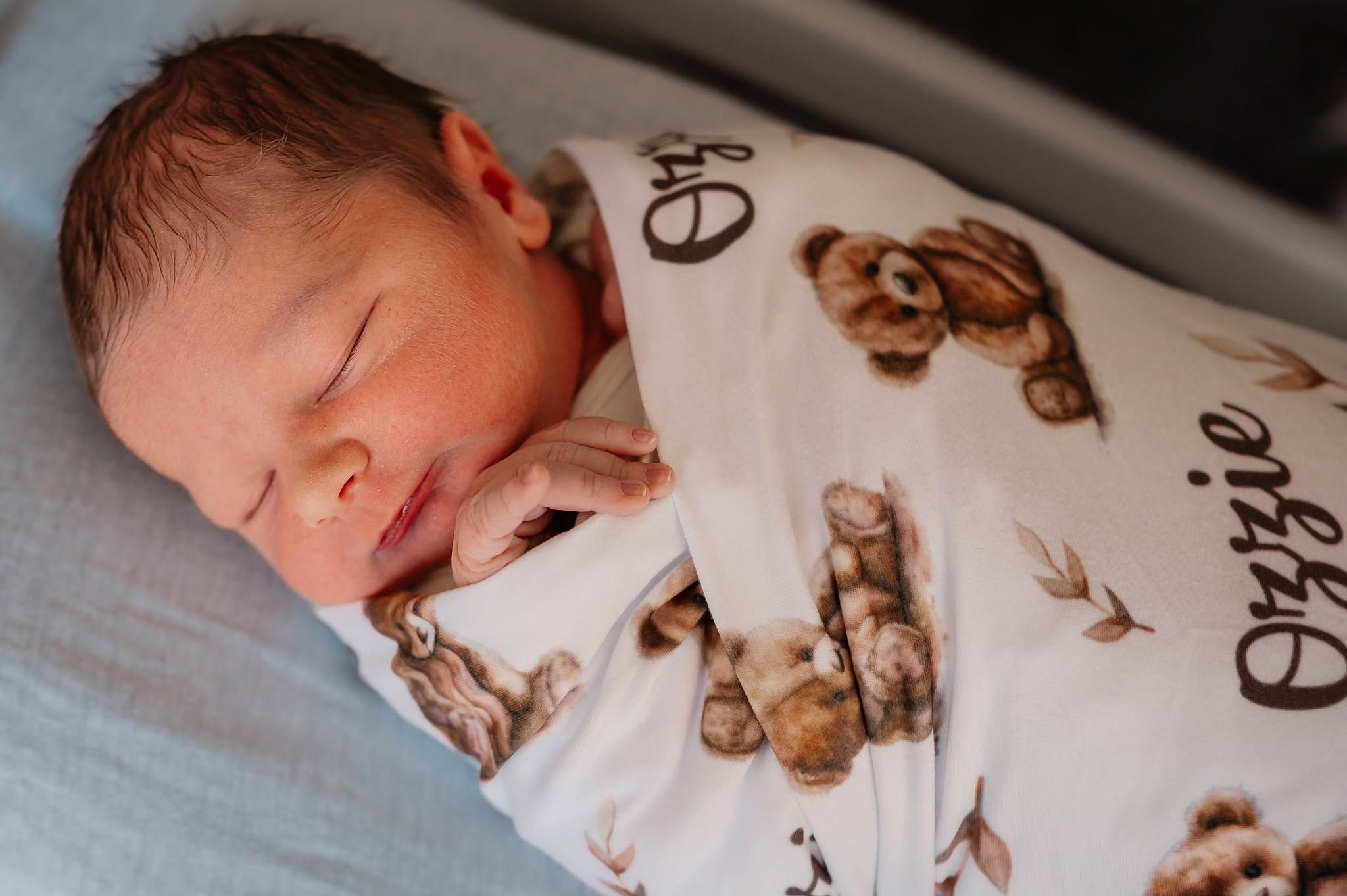 Sleeping newborn baby named Ozzie swaddled in a personalized teddy bear print hospital blanket.