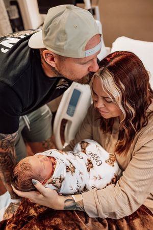 New parents gazing lovingly at their newborn baby wrapped in teddy bear swaddle in hospital room.