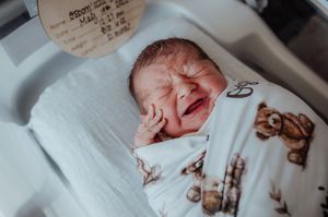 Crying newborn baby Ozzie swaddled in teddy bear print blanket in hospital bassinet after birth.
