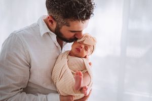 Father tenderly kisses sleeping newborn baby wrapped in cream swaddle.