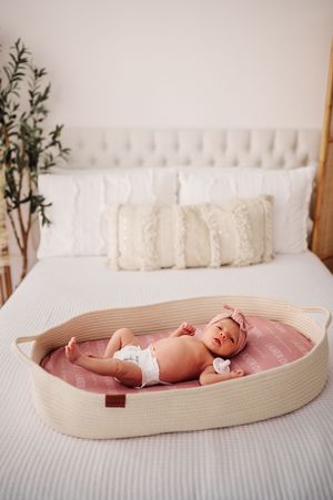 Newborn baby girl in diaper lying in rope moses basket on bed with pink personalized blanket.