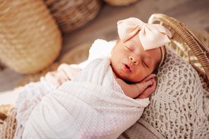 Sleeping newborn baby girl swaddled in white wrap with a pink bow headband in a wicker basket.
