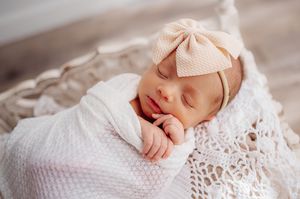 Sleeping newborn baby girl in pink bow headband wrapped in white blanket.