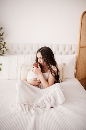 A mother with dark hair sits on a white bed cradling her newborn baby wrapped in a white blanket.