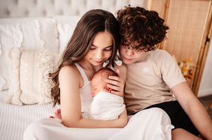 A young couple sits together on a bed tenderly holding their newborn baby wrapped in white.