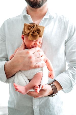 Father in white linen shirt tenderly holding sleeping newborn baby girl with brown bow headband.