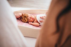 Newborn baby girl sleeping peacefully in a moses basket with a brown bow headband on a pink blanket.