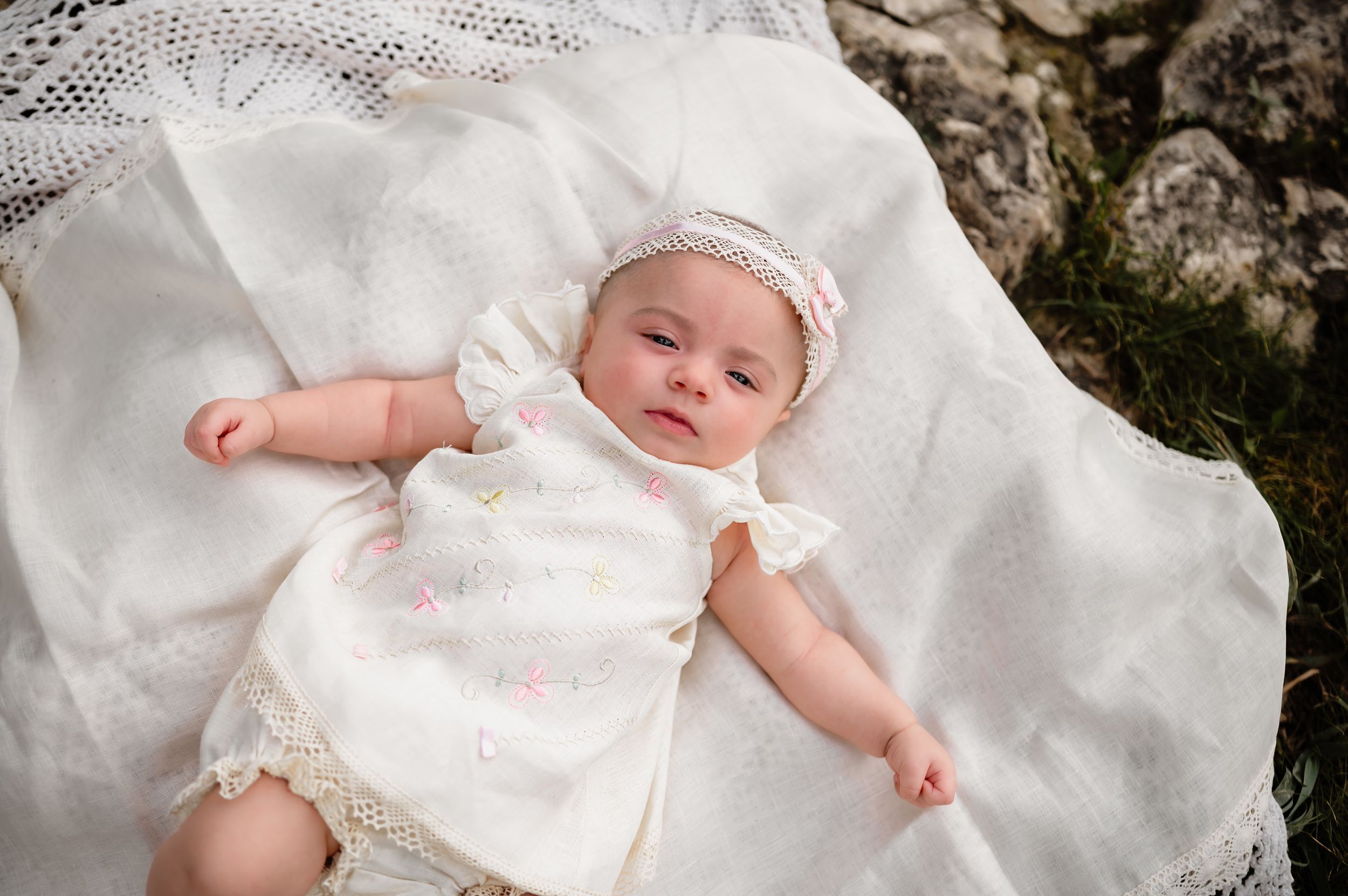 A newborn baby girl in a white floral dress and headband lies on a white lace blanket outdoors.