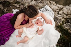 A mother in a purple dress leans down to kiss her newborn baby lying on a white lace blanket on rocks.