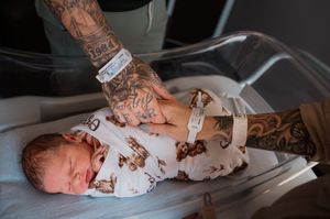 A tattooed parent gently holds a swaddled newborn baby in a hospital bassinet with a bear-print blanket.