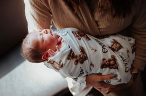 A mother in a tan sweater holds her yawning swaddled newborn baby wrapped in a bear-print blanket.