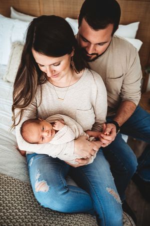 A couple in casual clothing sits near a bed lovingly gazing at their sleeping newborn baby.