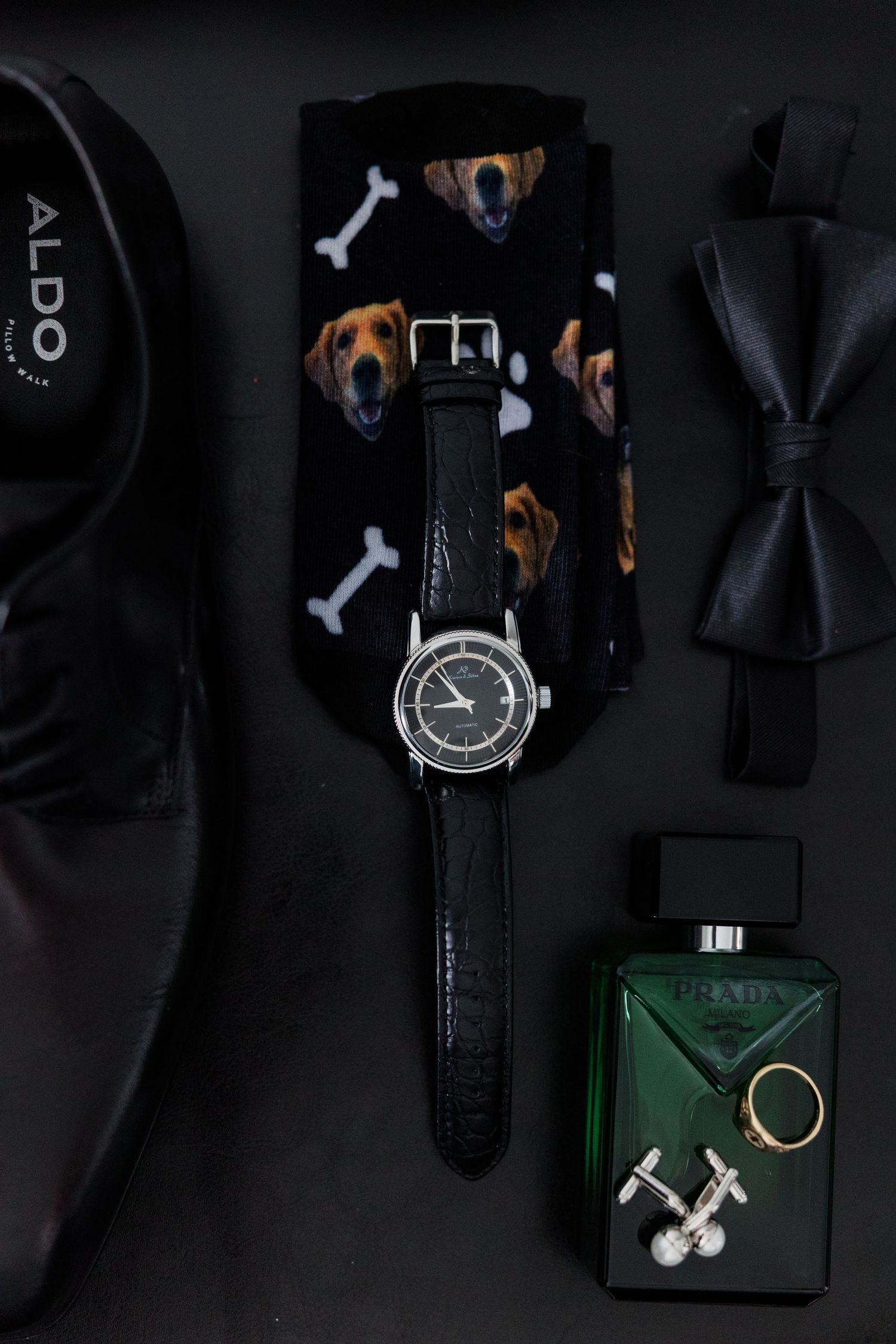 Men's wedding accessories flat lay featuring Aldo shoes, dog socks, watch, Prada cologne, cufflinks, gold ring, and black bow tie.