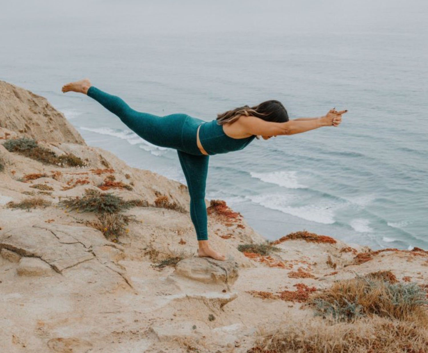 Woman practicing yoga warrior pose on rocky coastal cliffs in teal activewear with ocean waves in Newport Beach, California.