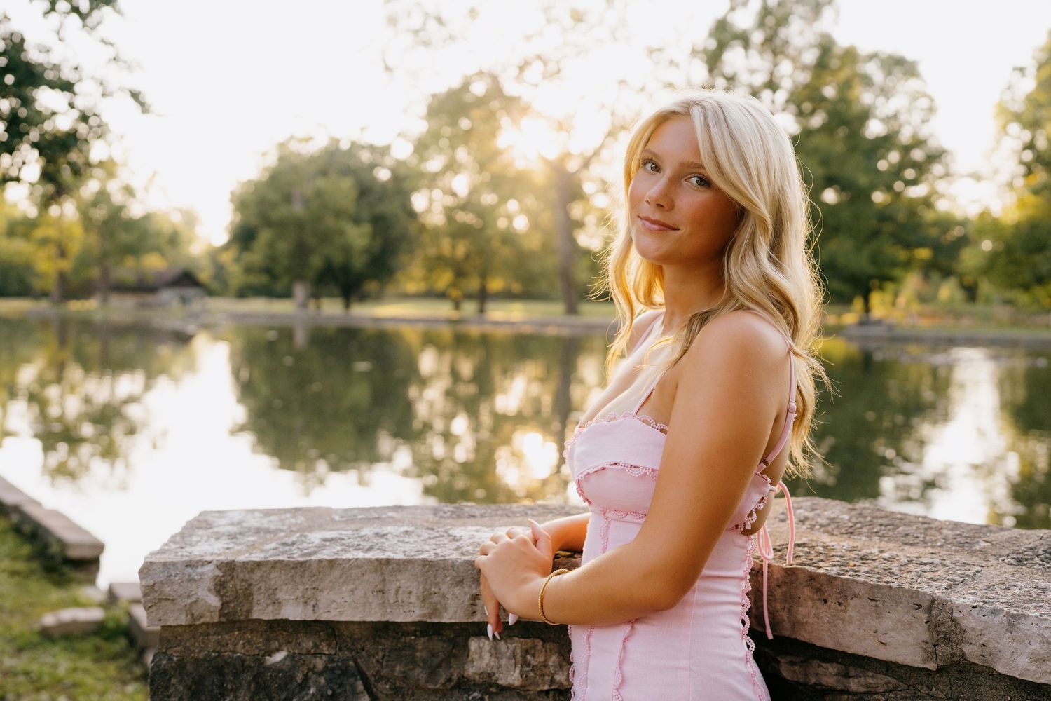 Blonde woman in pink dress leaning on stone wall by a sunlit pond in a lush green park.