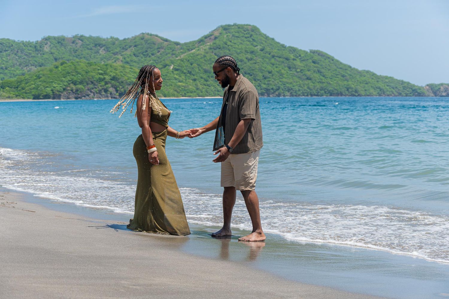 Couple holding hands on a tropical beach with turquoise water and lush green hills in the background.