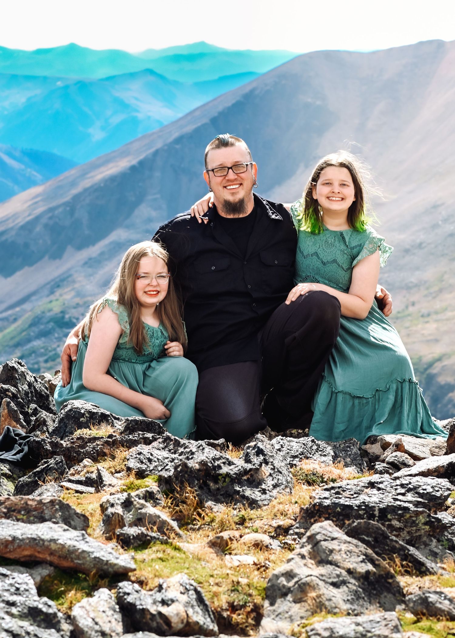 A man and two girls in matching teal dresses smile together on a rocky mountain summit with scenic blue peaks behind.