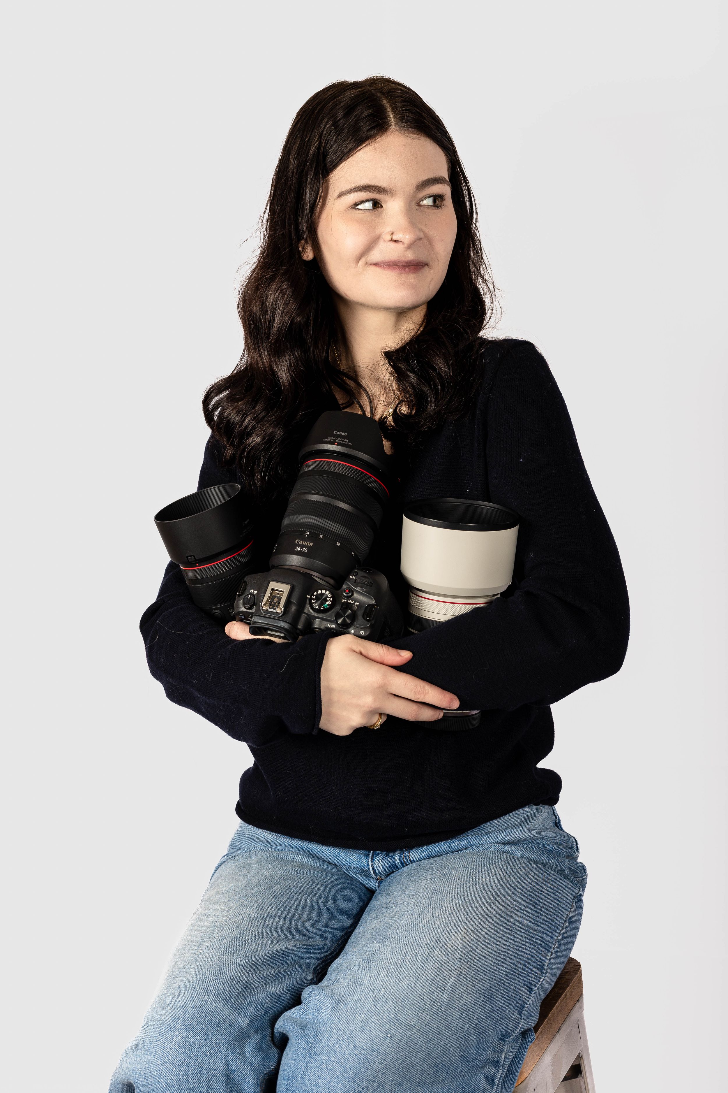 Young woman photographer sitting and holding multiple camera lenses and DSLR camera, wearing black sweater and jeans.