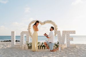 Man proposes to woman on beach with large LOVE letter signs and floral heart arch in background.
