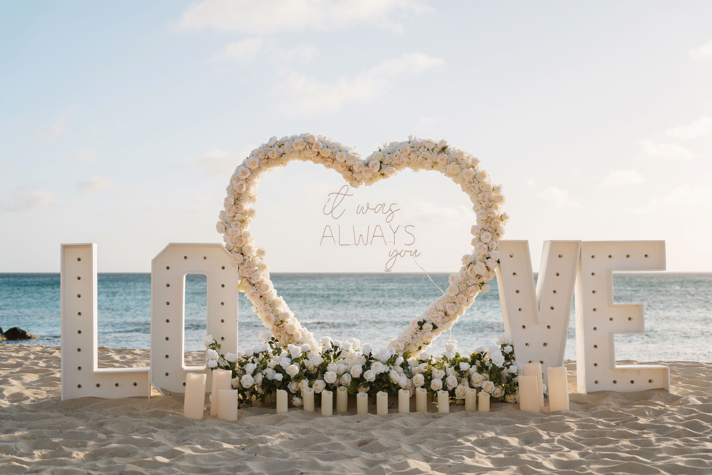 Beach wedding decor with large LOVE letters and a floral heart arch on sandy shore at sunset.