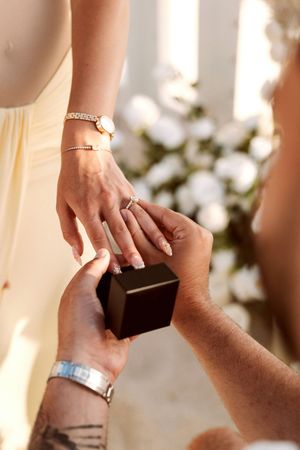 Man placing a diamond engagement ring on woman's finger, with a ring box, bracelets, and white flowers in background.