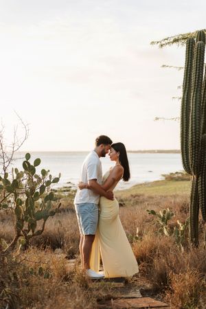 Couple embracing near cacti with ocean backdrop at golden hour in a romantic outdoor setting.