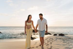 Couple holding hands walking on a tropical beach at sunset, woman in yellow dress, man in blue shorts.