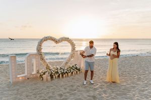 Couple on beach at sunset with floral heart arch and LOVE letters for a romantic proposal setup.