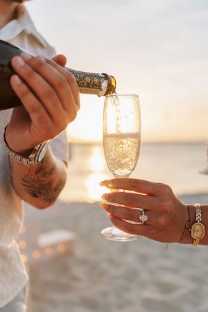 Couple celebrating engagement on beach at sunset, pouring champagne into flute, diamond ring visible on womans hand.