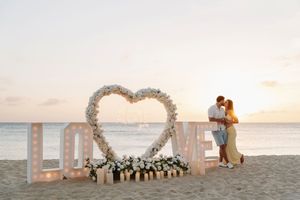 Couple kissing at sunset on beach beside floral heart arch and illuminated LOVE letters with white flowers.
