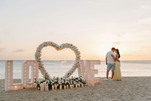 Couple kissing at sunset on beach beside illuminated LOVE sign with heart-shaped floral arch and white flowers.