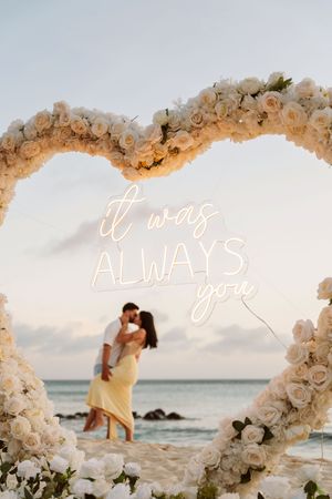 Couple kissing on beach framed by heart-shaped white rose arch with neon sign reading it was always you.