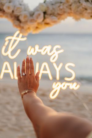Hand with engagement ring raised near neon sign reading 'it was always you' on a beach with floral arch.