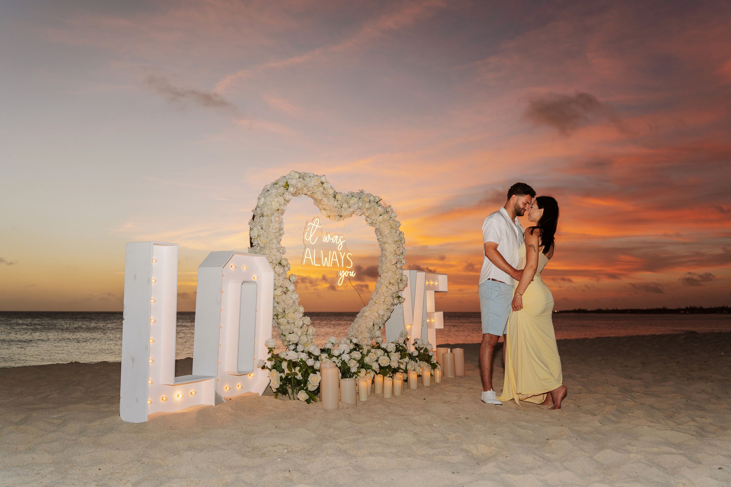 Couple kissing at sunset beach proposal with floral heart arch, LOVE letters, and candles in Aruba.