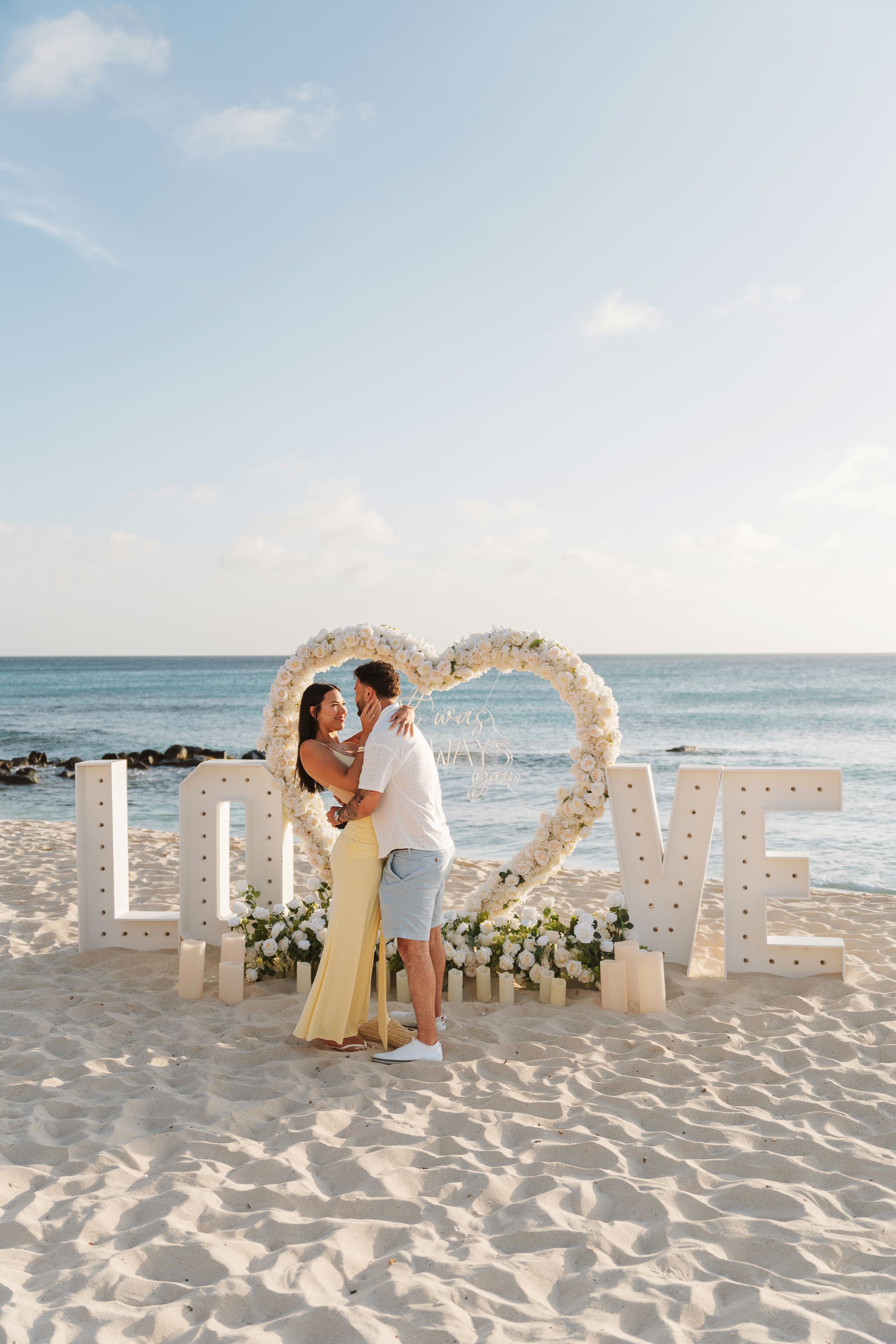 Couple kissing in front of a floral heart arch and LOVE letters on a tropical beach at sunset.
