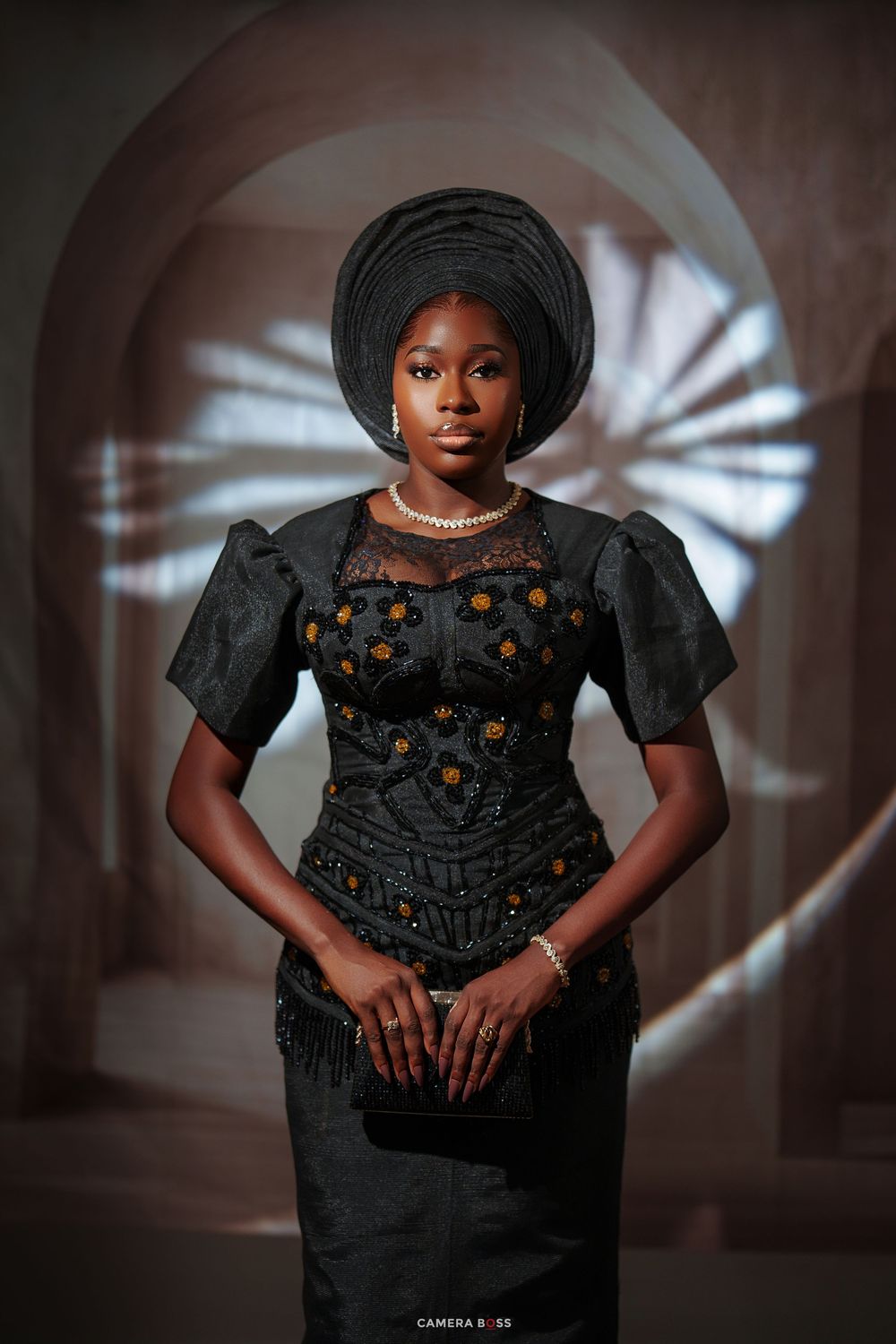 Woman in black embroidered Nigerian traditional attire with gele headwrap and pearl necklace posing elegantly.