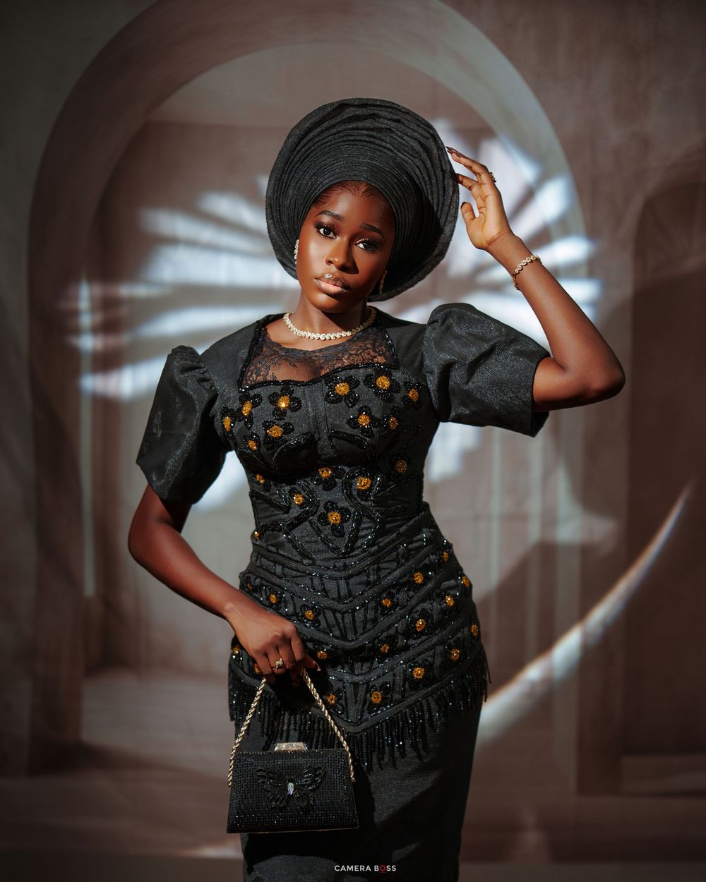 Woman in black embroidered dress and gele headwrap holding handbag in elegant Nigerian traditional attire.