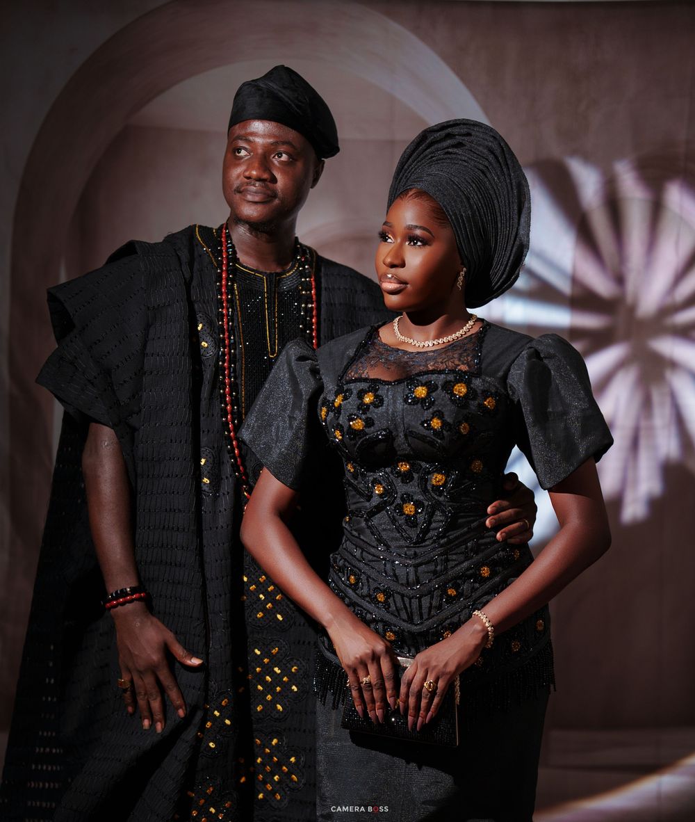 Nigerian couple in traditional Yoruba attire, man in black agbada with cap, woman in embroidered black dress with gele headwrap.